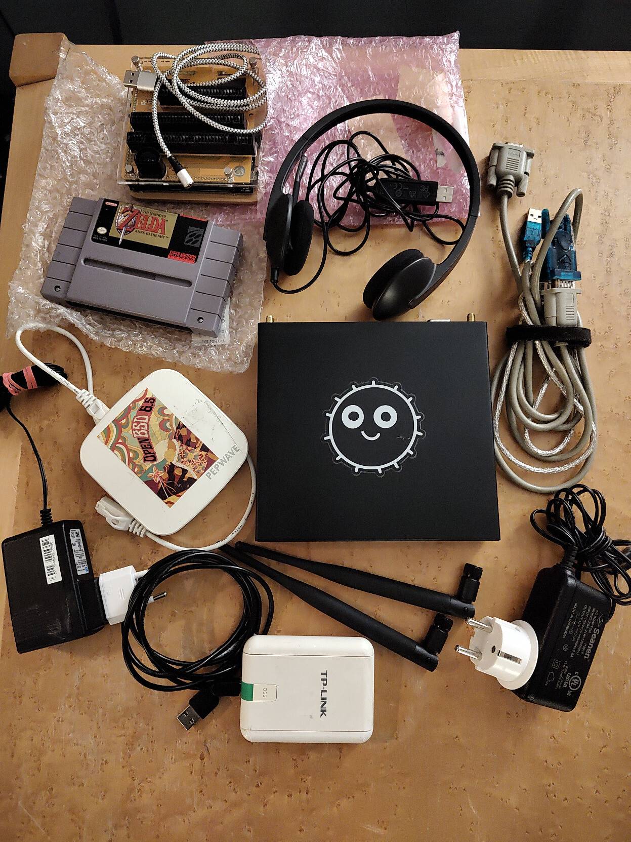 Various cables and devices lying on a wooden table. The devices are a black APU at the center, a black USB audio headset towards the top, a small and white wireless access point to the left, a small and white wireless USB device at the bottom, a SNES cartridge of Zelda 3 and a cardtridge reader in the upper left corner. Also visible are two unscrewed wifi antennas and two power plugs.