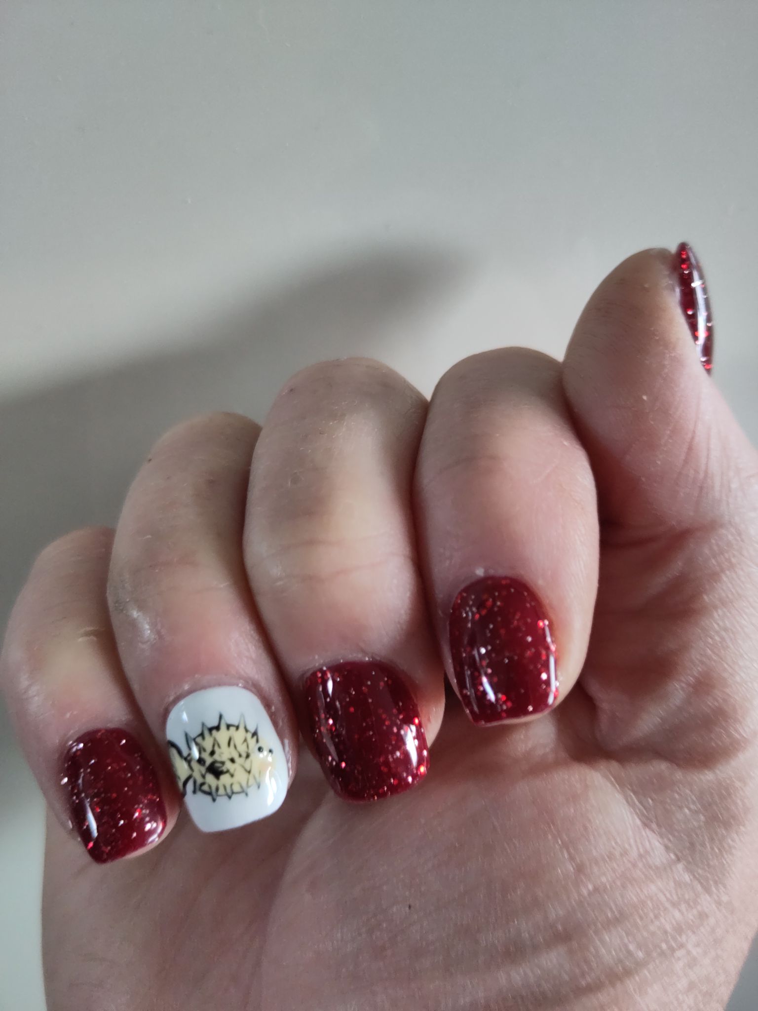 A photo of my acrylic finger nails painted i n a red glitter and on my ring finger a white background with a pufferfish painted on aka the OpenBSD logo.