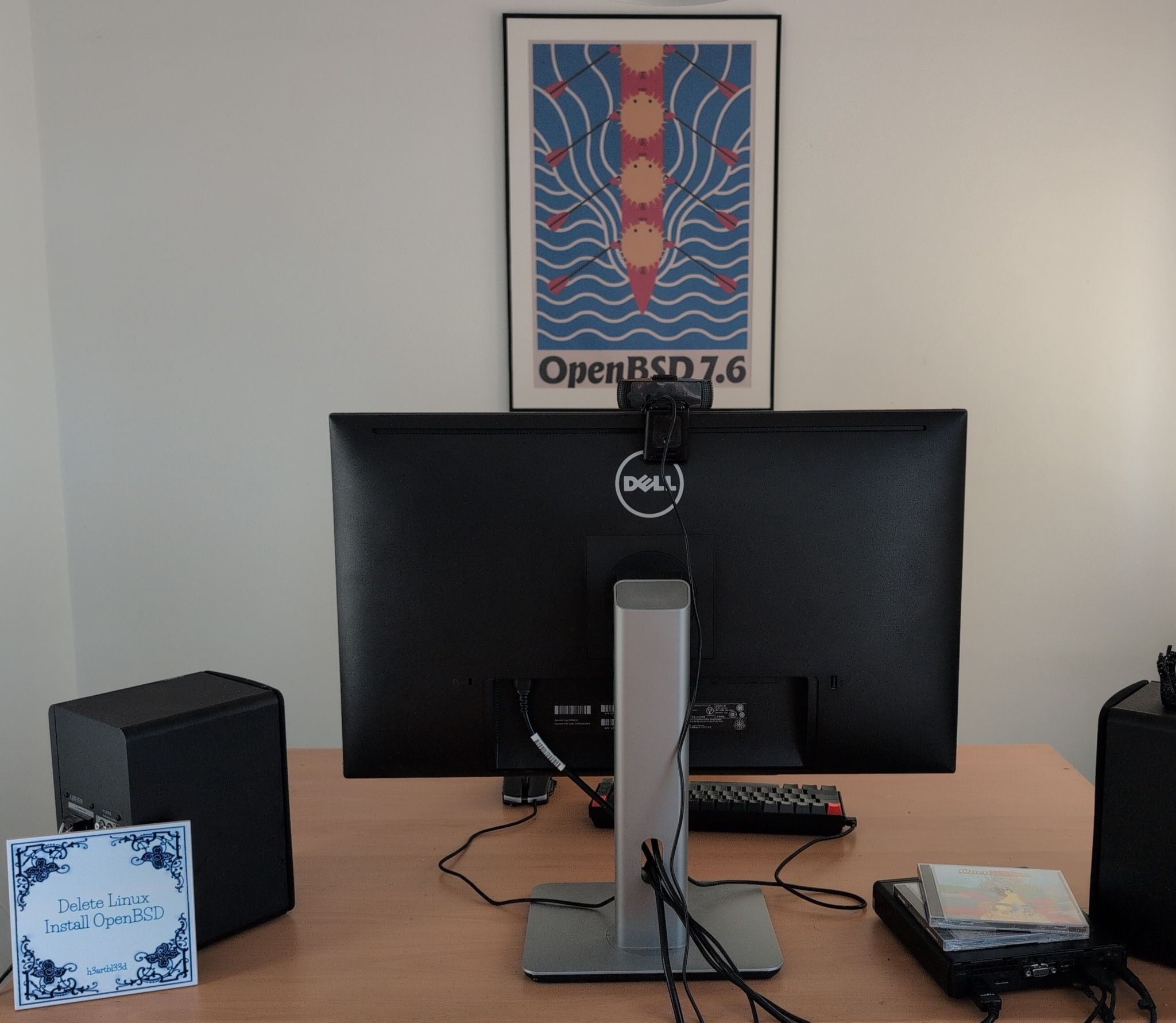 Photo showing a desktop, with the OpenBSD 7.6 release art hanging on the wall, a tile with "delete Linux install OpenBSD" and the release of 3.7 laying on the mini PC. There is a large monitor visible and keyboard, mouse, and two speakers.