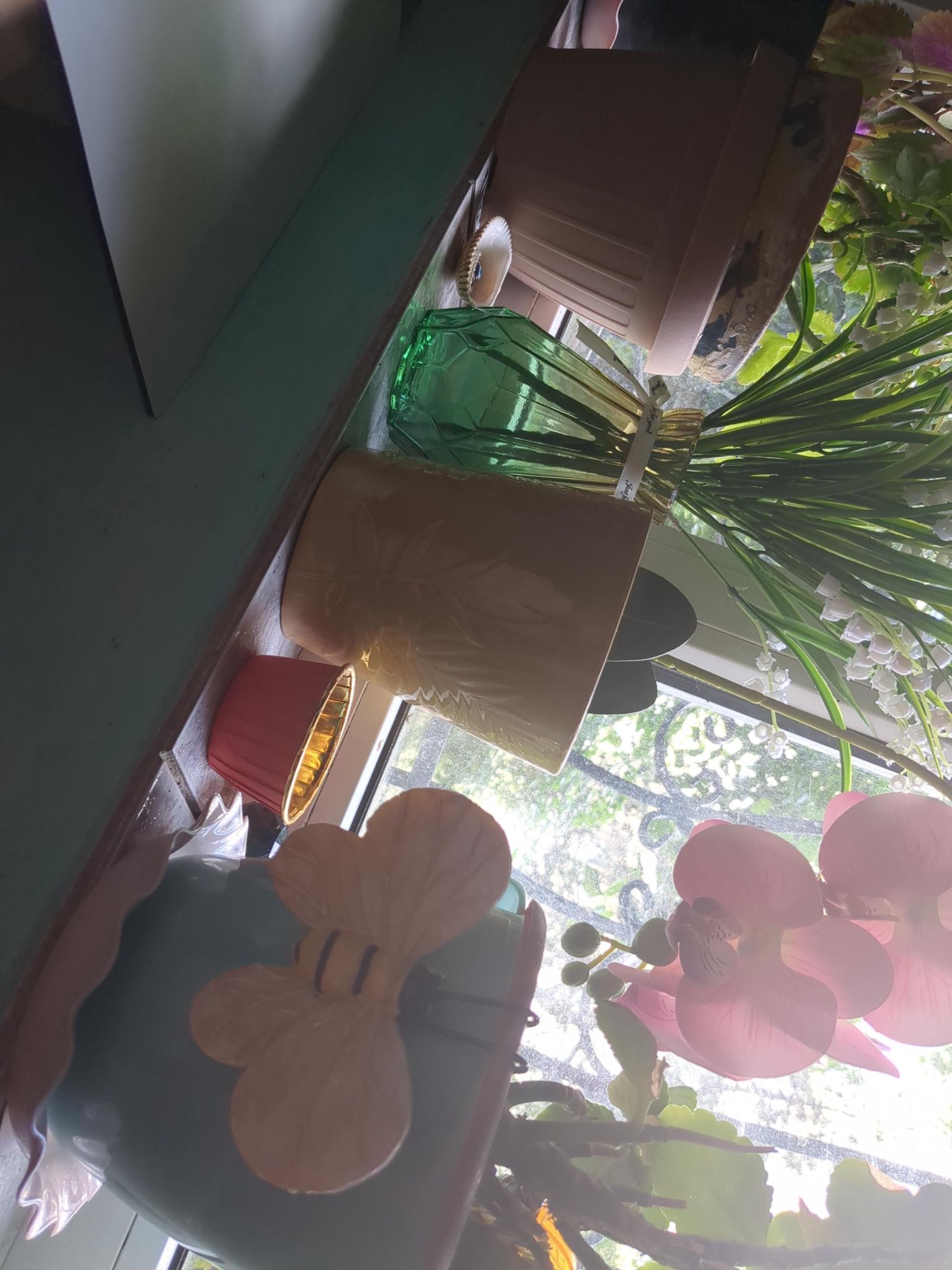 Colorful real and artificial plants in fancy vases, decorated with fake bees with anthropomorphic eyes hanging to the vases... Windowsill, outside the window there's also greenery.